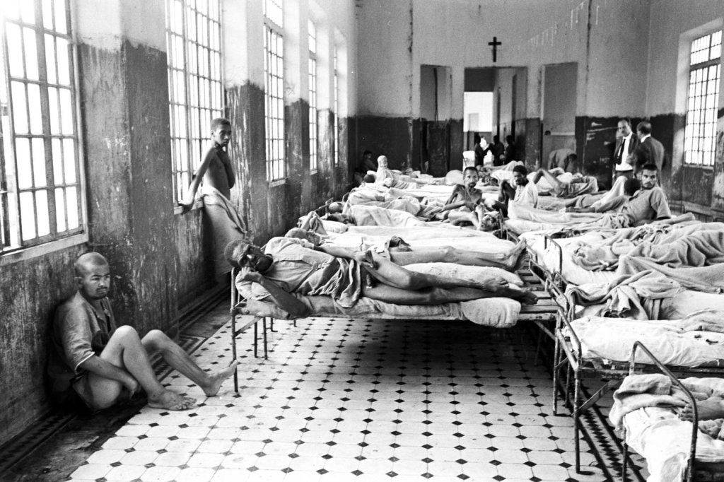 Homens em um hospital superlotado, alguns deitados em camas simples, outros sentados no chão ou em parapeitos de janelas. Um homem magro olha para a câmera, enquanto outros parecem doentes ou exaustos. O ambiente é precário, com paredes descascadas e janelas gradeadas. Ao fundo, uma cruz na parede e dois homens de terno observam a cena. A imagem em preto e branco transmite uma atmosfera de sofrimento e abandono.