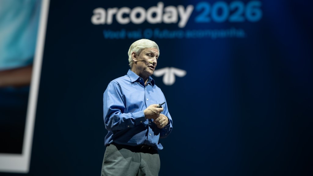 Homem idoso de cabelos brancos, camisa azul e calça cinza, falando em um palco escuro com um microfone na mão. Ao fundo, um telão exibe