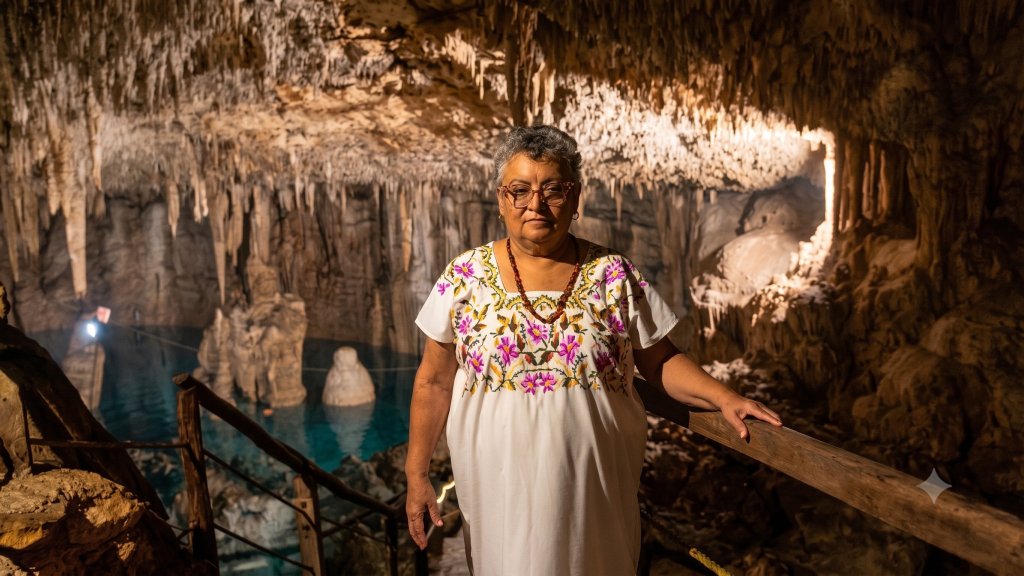 Mulher de pele clara e cabelos grisalhos curtos, usando óculos e um vestido branco bordado com flores roxas e rosas, segura um corrimão de madeira dentro de uma caverna iluminada. Ao fundo, estalactites e estalagmites formam um cenário rochoso, com um lago de água azul-turquesa visível à esquerda
