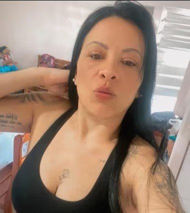 Mulher de cabelos pretos longos, com tatuagens nos braços e no peito, vestindo top preto, fazendo bico para a câmera em um ambiente interno
