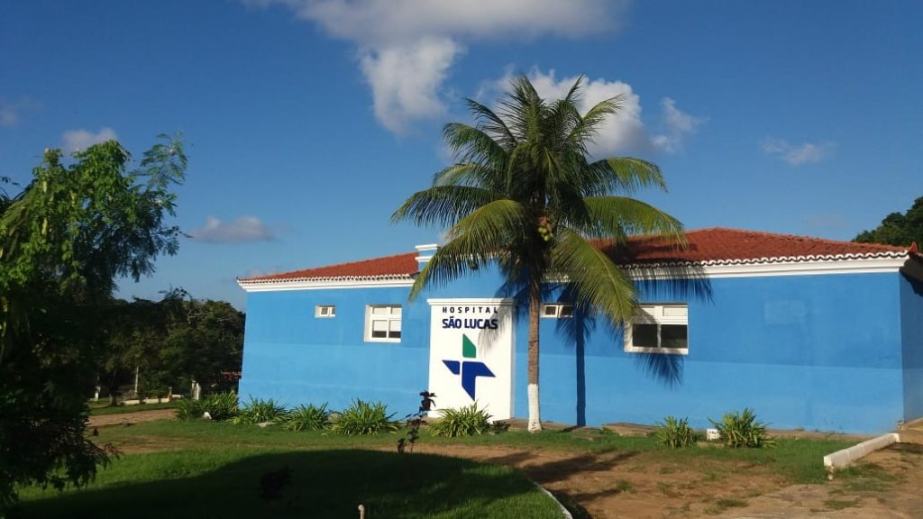 Fachada do Hospital São Lucas, um prédio azul com telhado de cerâmica laranja, sob um céu azul com nuvens esparsas. Em frente, um coqueiro alto e um letreiro branco com o nome do hospital e um logotipo azul e verde. Há gramado e vegetação ao redor