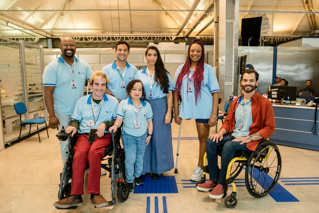 Grupo de sete pessoas sorrindo, vestindo camisetas azuis claras com crachás. Dois homens em cadeiras de rodas, uma mulher com muleta, uma mulher de baixa estatura, e três pessoas em pé. Ao fundo, um ambiente de escritório com divisórias e mesas. No chão, piso tátil azul.