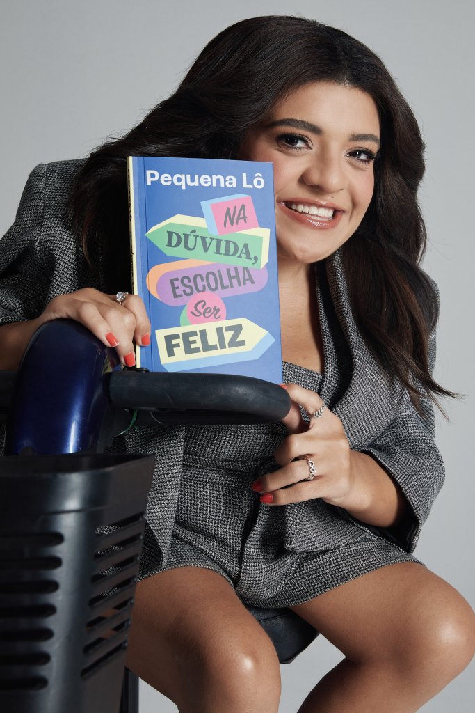 Pequena Lô sorrindo, segurando um livro azul com o título Pequena Lô: Na Dúvida, Escolha Ser Feliz. Ela veste um blazer e shorts xadrez cinza, com unhas vermelhas e anéis prateados, sentada em uma cadeira de rodas azul e preta